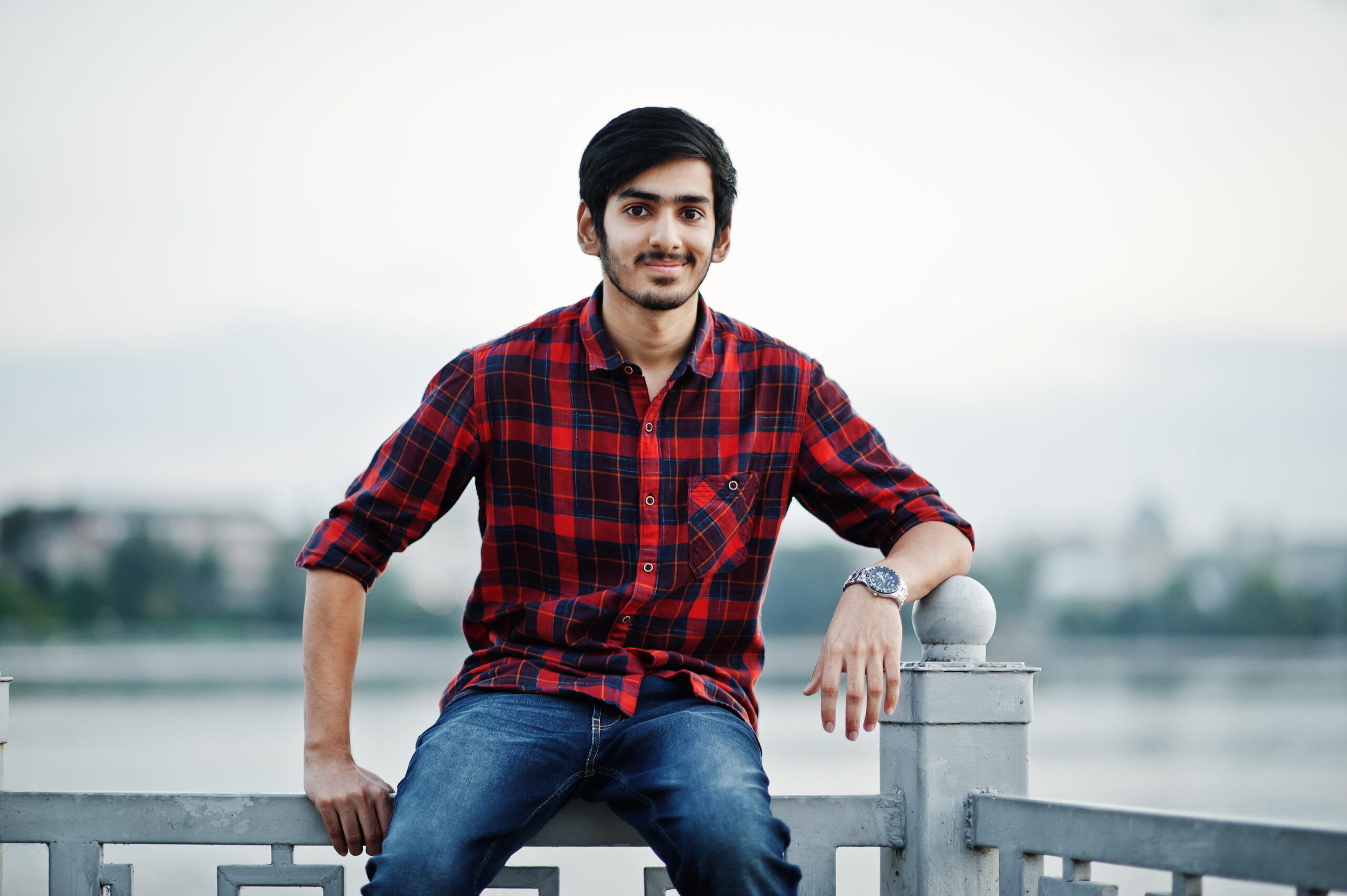 young indian student man checkered shirt jeans sitting handrails against lake scaled